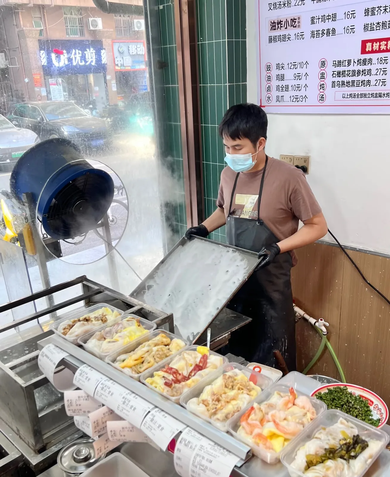 Cook pulling a tray from a drawer steamer at a cheung fun shop, with trays of rice noodle rolls ready on the counter
