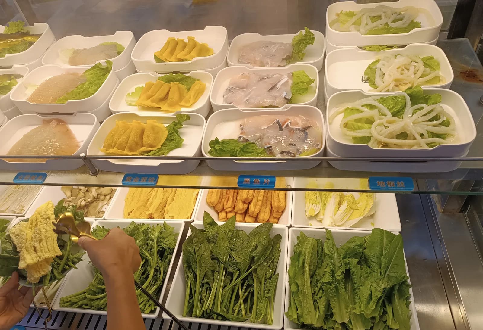 Self-serve counter at a Shenzhen hot pot buffet with trays of leafy greens, tofu skin and sliced fish