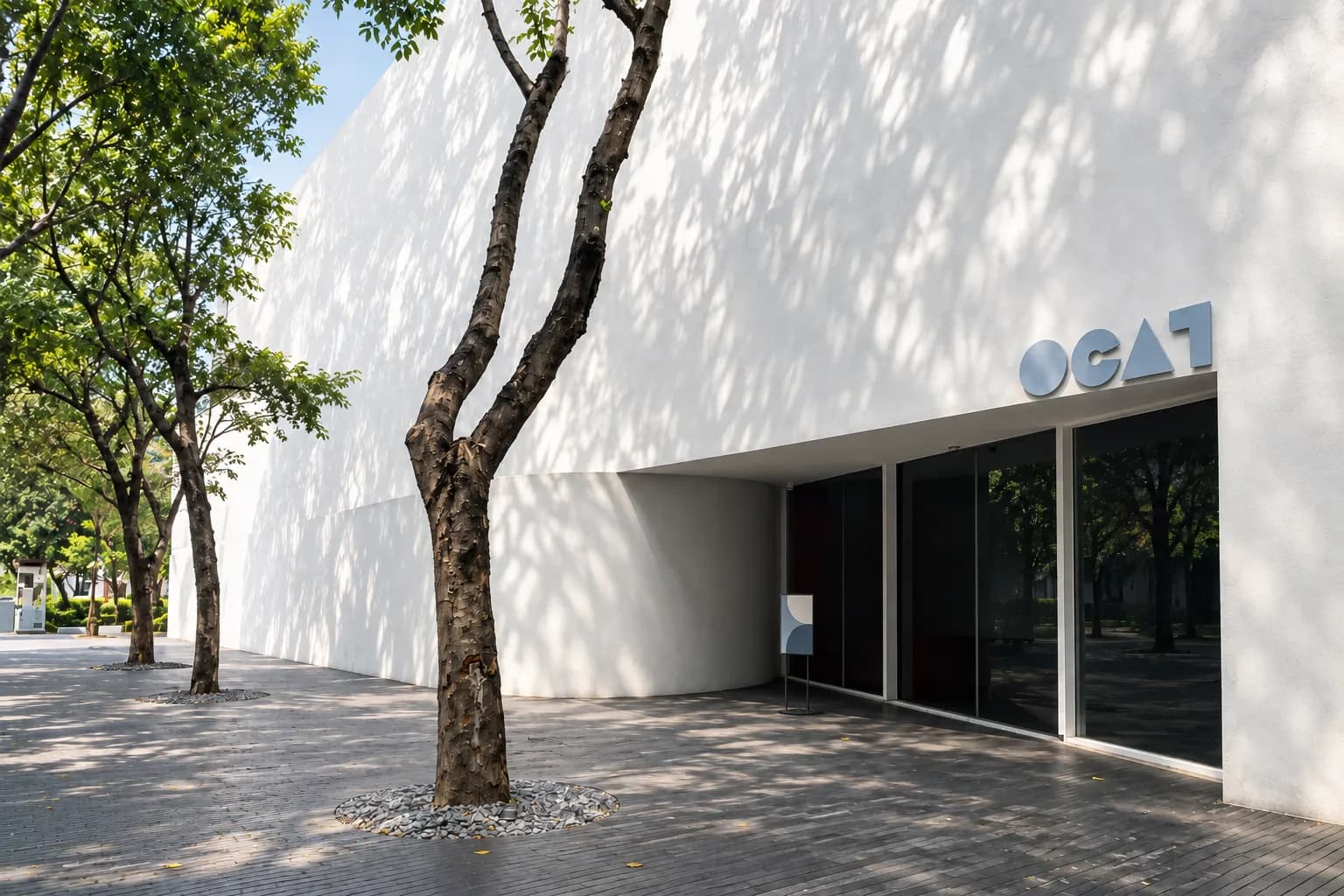 Curved white wall of OCAT Shenzhen with the blue OCAT sign above its glass doors, beside a tree-lined walkway