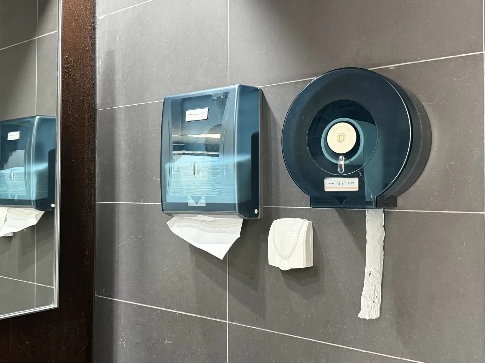A shared jumbo toilet roll dispenser and a paper towel dispenser on the wall outside the stalls in a public restroom